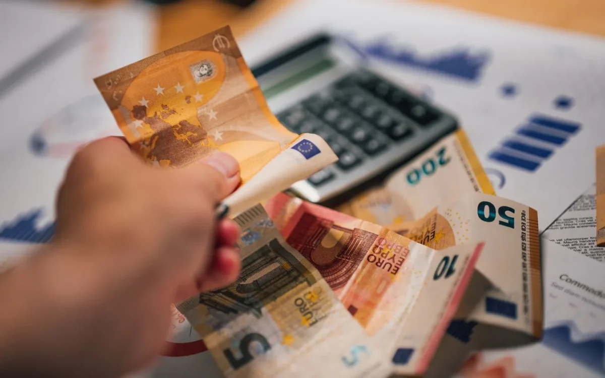 Mână cu bancnote euro deasupra unor documente și a unui calculator