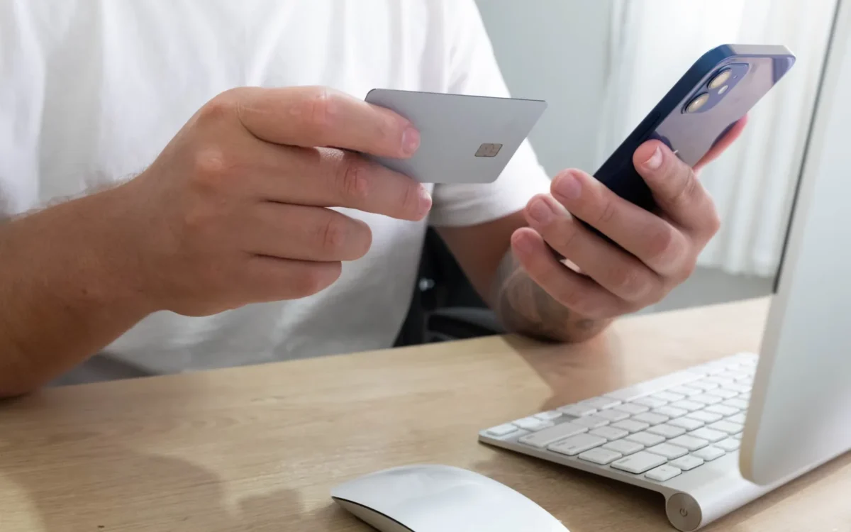 Persoană ținând un card bancar și un telefon mobil lângă calculator