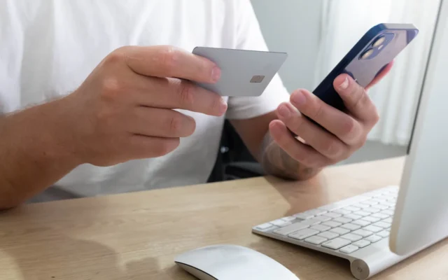 Persoană ținând un card bancar și un telefon mobil lângă calculator