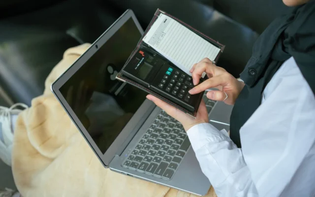 Mâini care folosesc un calculator de buzunar lângă un laptop deschis