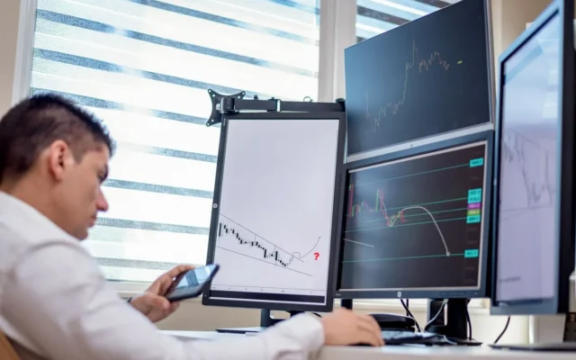 Persoană cu telefon în mână, în fața unor monitoare cu grafice financiare