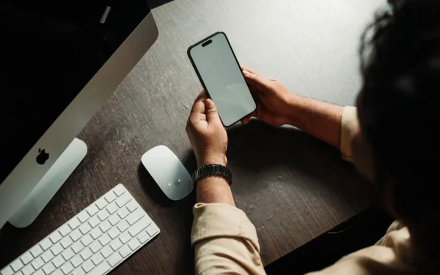 Persoană care ține un telefon mobil lângă un computer.
